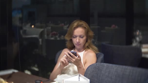 Красива жінка з телефоном ввечері чекає в ресторані. 4k, розмивання тла — стокове відео