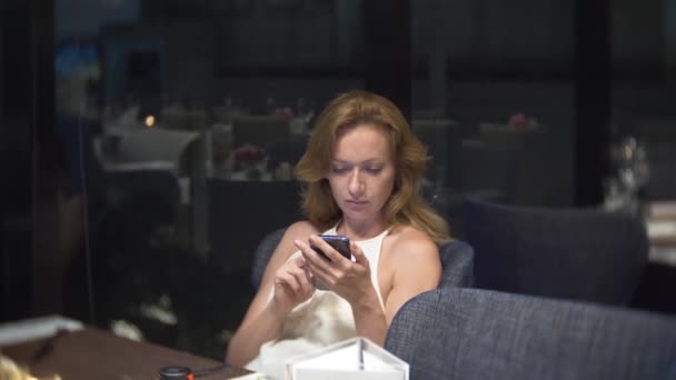 Hermosa mujer con un teléfono por la noche esperando en el restaurante. 4k, desenfoque de fondo — Vídeos de Stock