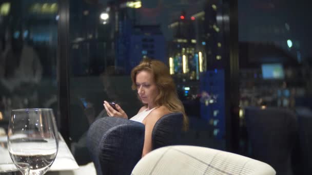 Hermosa mujer con un teléfono por la noche esperando en el restaurante. 4k, desenfoque de fondo — Vídeo de stock