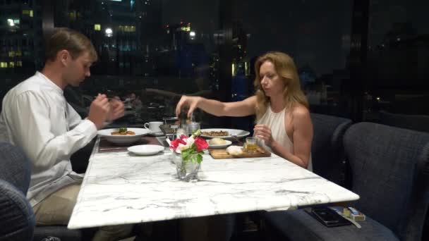 Счастливая пара разговаривает и ужинает в баре с видом на небоскребы, вечером, 4k, размытый фон — стоковое видео
