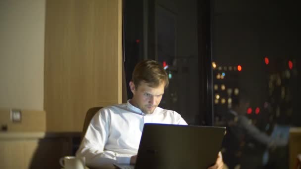 Un hombre joven y guapo usando un portátil en una silla en una habitación con una ventana panorámica con vistas a los rascacielos por la noche. 4k, borra el fondo . — Vídeo de stock