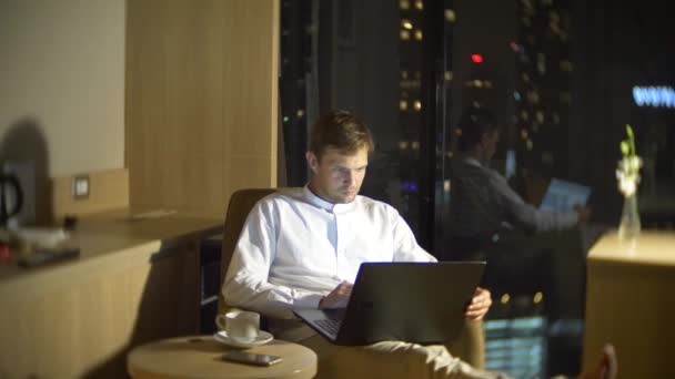 一个年轻英俊的男子, 用笔记本电脑在一个房间的椅子上, 在一个全景窗口俯瞰摩天大楼在夜间。4k、模糊背景. — 图库视频影像