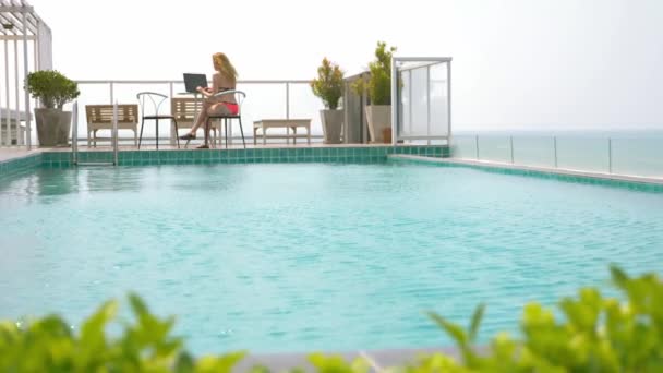 Una mujer independiente sentada junto a la piscina en el techo con vistas al océano y trabajando en un ordenador portátil al atardecer. 4K, cámara lenta — Vídeos de Stock