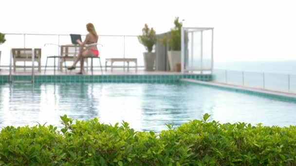 Женщина-фрилансер, сидящая у бассейна на крыше с видом на океан и работающая на ноутбуке на закате. 4К, замедленная съемка — стоковое видео