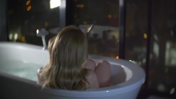 Bella donna che si gode una rilassante vasca da bagno in un lussuoso bagno con finestra di notte. Il concetto di uno stile di vita e di bellezza. vista dalla finestra ai grattacieli. 4k — Video Stock