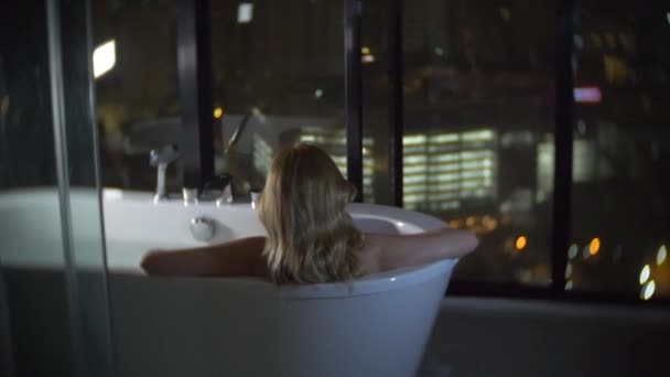 Schöne Frau genießt ein entspannendes Bad in einem luxuriösen Badezimmer mit Fenster in der Nacht. das Konzept einer Lebensweise und Schönheit. Blick vom Fenster auf die Wolkenkratzer. 4k — Stockvideo