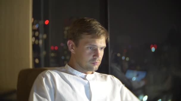 Un jeune et beau homme utilisant un ordinateur portable sur une chaise dans une chambre avec une fenêtre panoramique donnant sur les gratte-ciel la nuit. 4k, brouiller le fond . — Video