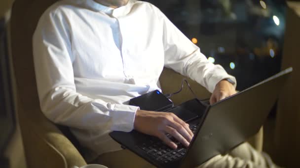 Een jonge, knappe man met een laptop op een stoel in een kamer met een panoramisch venster met uitzicht op de wolkenkrabbers in de nacht. 4k, wazig de achtergrond. — Stockvideo