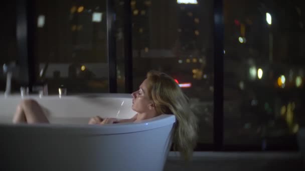 Krásná žena se těší relaxační koupel v luxusní koupelnu s oknem v noci. Koncept způsobu života a krásy. pohled z okna na mrakodrapy. 4k