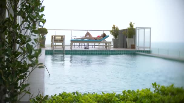 Eine Frau im rosafarbenen Bikini genießt den Blick auf das Meer am Pool auf dem Dach, das Mädchen sonnt sich auf einer Liege. 4k, Zeitlupe — Stockvideo