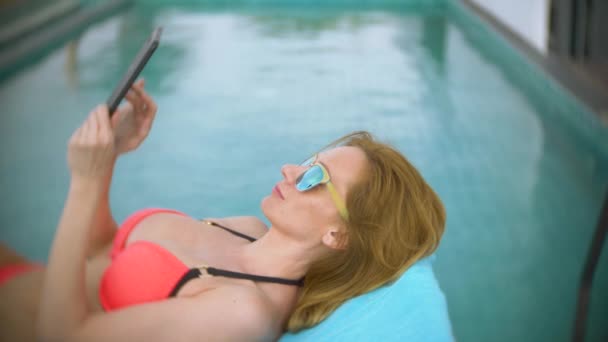 Una mujer en bikini rosa junto a la piscina en el techo, una chica tomando el sol en una tumbona y usando el teléfono. 4k, cámara lenta — Vídeo de stock