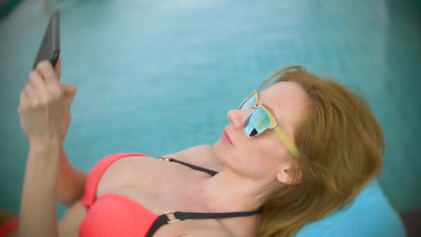 Uma mulher de biquíni rosa junto à piscina no telhado, uma menina tomando banho de sol em uma cadeira de praia e usando o telefone. 4k, câmera lenta — Vídeo de Stock