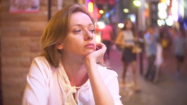 Mujer joven y hermosa sentada en la calle en un café o terraza. Una mujer observa a los transeúntes caminando por una calle peatonal por la noche. 4k . — Vídeos de Stock