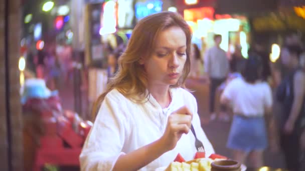 Mujer joven y hermosa comiendo postre, sentada afuera en una cafetería o terraza. Una mujer admira un corte de fruta. 4k . — Vídeo de stock