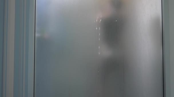 Mulher atrás de vidro desfocado. Menina se preparando tomar banho. Uma mulher na casa de banho. um homem observa como uma mulher toma um banho através de uma parede de vidro no chuveiro. 4k — Vídeo de Stock