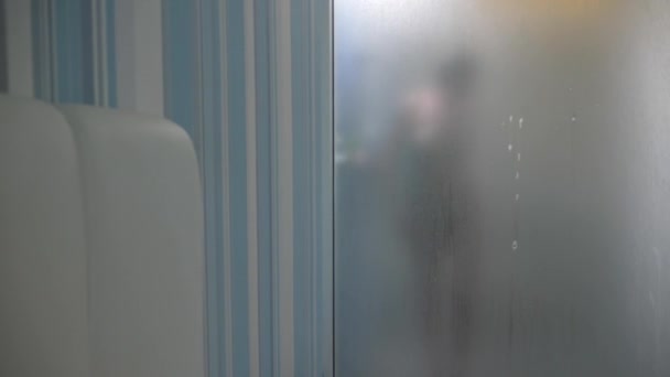 ぼやけたガラスの背後にある女性。準備中の女の子は、シャワーを浴びる。浴室で女性。男の時計として女性シャワーのガラスの壁のシャワーがかかり。4 k — ストック動画