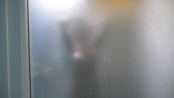 Žena za rozmazaná sklem. Dívka, připravuje si sprchu. Žena v koupelně. muž hodinky, jako žena bere sprchu přes skleněnou stěnu ve sprše. 4k — Stock video