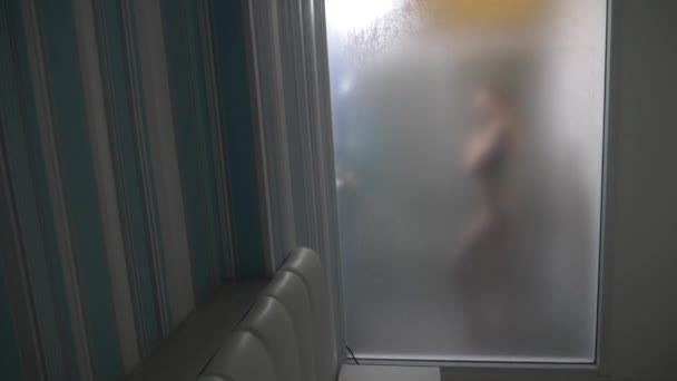 Mujer detrás de vidrio borroso. Chica preparando tomar ducha. Mujer en el baño. un hombre observa como una mujer toma una ducha a través de una pared de vidrio en la ducha. 4k — Vídeos de Stock