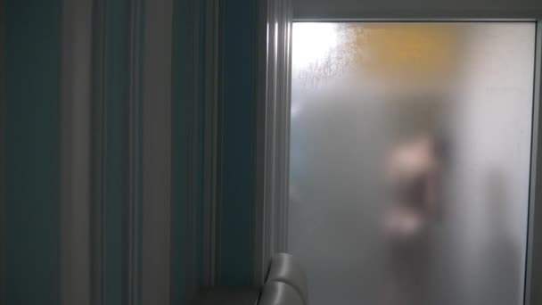 Mujer detrás de vidrio borroso. Chica preparando tomar ducha. Mujer en el baño. un hombre observa como una mujer toma una ducha a través de una pared de vidrio en la ducha. 4k — Vídeos de Stock