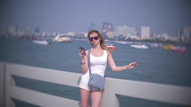 Женщина в солнечных очках, использует свой смартфон, стоя на пирсе. 4K, размытие фона, наклон вниз — стоковое видео