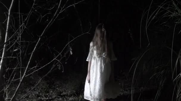 Ein kleines Geistermädchen mit langen schwarzen Haaren, in weiß, wandert mit einem Messer und einem Stofftier durch den Wald. 4k — Stockvideo
