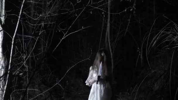 Una niña fantasma con el pelo largo y negro, en blanco, vagando por el bosque con un cuchillo y un juguete suave. 4k — Vídeos de Stock