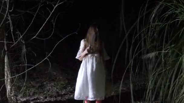 Una niña fantasma con el pelo largo y negro, en blanco, vagando por el bosque con un cuchillo y un juguete suave. 4k — Vídeo de stock