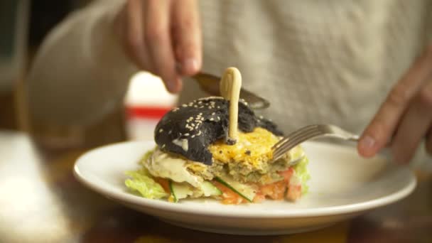 Ο άνθρωπος σε ένα λευκό πουλόβερ που τρώει ένα μαύρο μπέργκερ σε ένα εστιατόριο. 4k — Αρχείο Βίντεο