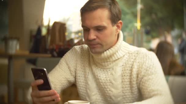 Knappe jonge man in een witte trui met behulp van een smartphone, zittend in een café in de stad op een tegen een raam. achtergrond vervagen. 4k — Stockvideo