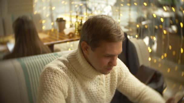Przystojny, młody człowiek w białym swetrze za pomocą smartfona, siedząc w kawiarni w mieście na przeciwko okna. Rozmycie tła. 4k — Wideo stockowe