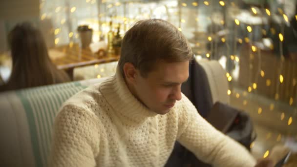 स्मार्टफोन वापरुन पांढ white्या स्वेटरमध्ये सुंदर तरुण माणूस खिडकीच्या विरूद्ध शहरातील एका कॅफेमध्ये बसला. पार्श्वभूमी अंधुक. 4k — स्टॉक व्हिडिओ