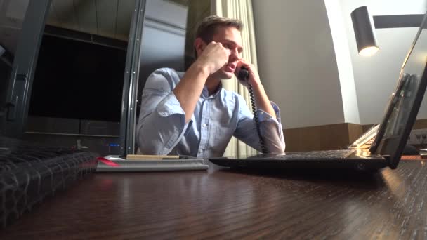 Jovem empresário falando ao telefone fixo, sentado à mesa no escritório à noite. caderno da frente. 4k — Vídeo de Stock