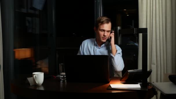 Fiatal üzletember beszél a vezetékes telefon, íróasztal hivatalban, az éjszaka ül. első jegyzetfüzet. 4k — Stock videók