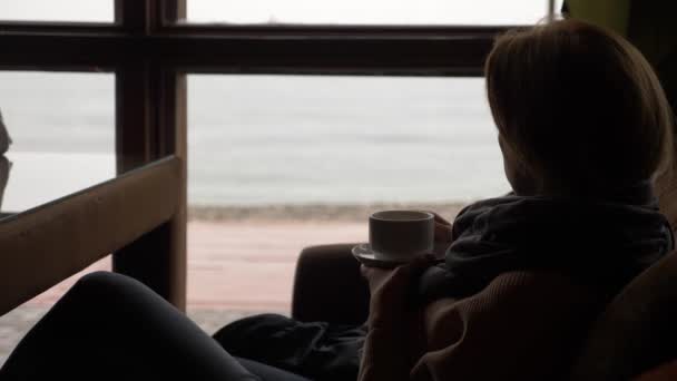 Mujer joven bebiendo té en la cafetería cerca de la ventana con vista al mar. una mujer en una bufanda y suéter está bebiendo té y tomando el sol en un café. 4k — Vídeos de Stock