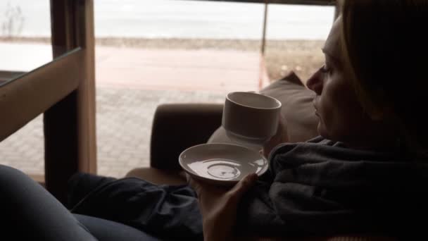 Jonge vrouw drinken van thee in café in de buurt van venster met uitzicht op zee. een vrouw in een sjaal en trui is het drinken van thee en koesteren in een café. 4k — Stockvideo