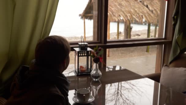Юнак п'є чай у кафе біля вікна з видом на море. Чоловік в шарфі і светрі п'є чай і занурюється в кафе. 4k — стокове відео