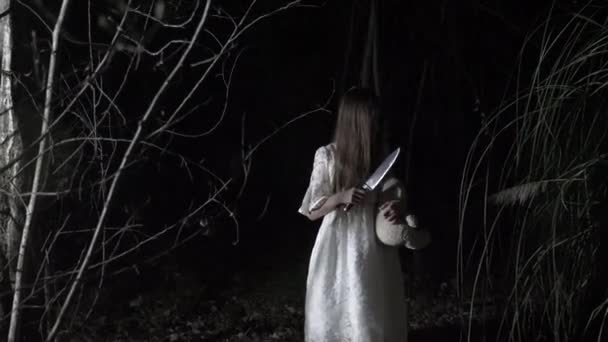 Een spook meisje met lange zwarte haren, in wit, zwerven door het bos met een mes en een zacht stuk speelgoed. 4k — Stockvideo