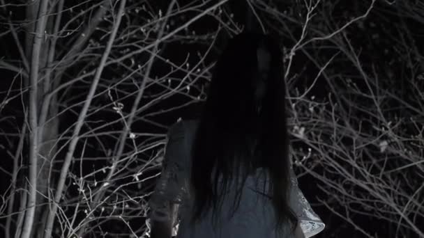 Een spook meisje met lange zwarte haren, in wit, zwerven door het bos met een mes en een zacht stuk speelgoed. 4k — Stockvideo