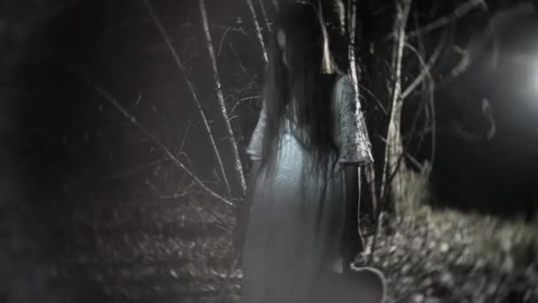 Маленькая девочка-призрак с длинными черными волосами, в белом, бродит по лесу с ножом и мягкой игрушкой. 4k — стоковое видео