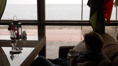 Genç kadın pencere Deniz Manzaralı yakınındaki kafede çay içme. bir kadın bir eşarp ve kazak çay içme ve bir kafede tadını çıkarma. 4k