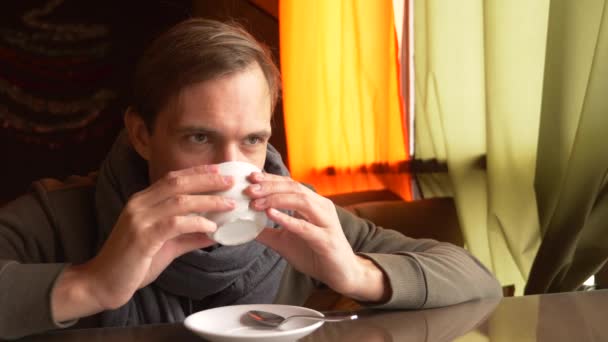 Joven con una bufanda bebiendo té y sonriendo en un café. 4k — Vídeo de stock