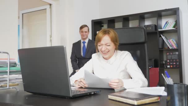 Два офисных работника сидят за столом, женщина работает за компьютером, рядом мужчина. Они смеются. 4k — стоковое видео