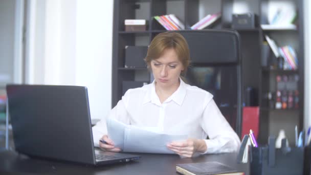 Женщина работает в офисе с документами и ноутбуком. она смотрит на камеру и улыбается 4k . — стоковое видео