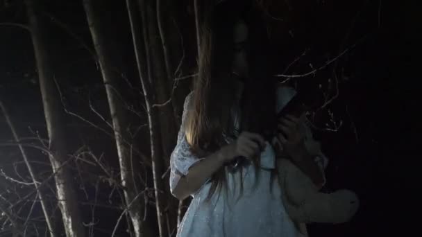 Una niña fantasma con el pelo largo y negro, en blanco, vagando por el bosque con un cuchillo y un juguete suave. 4k — Vídeo de stock