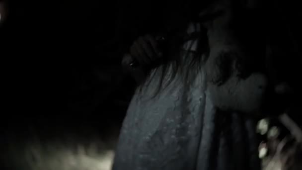 Маленька дівчинка-привид з довгим чорним волоссям, в білому кольорі, блукає лісом з ножем і м'якою іграшкою. 4k — стокове відео