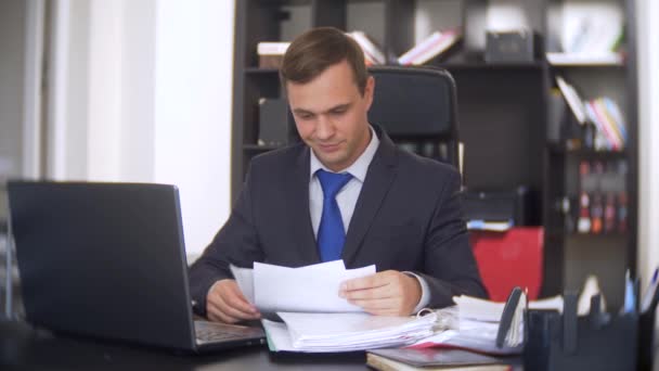 Hombre que trabaja en la oficina con documentos y portátil. mira a la cámara y sonríe 4k. muestra el pulgar hacia arriba — Vídeos de Stock