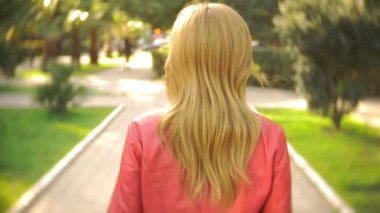 kamera sarışın kız izler. bir kadın bir kırmızı ceketli şehir parkı ile açık güneşli bir günde elini kolunu sallaya dönüyor. 4k, ağır çekim