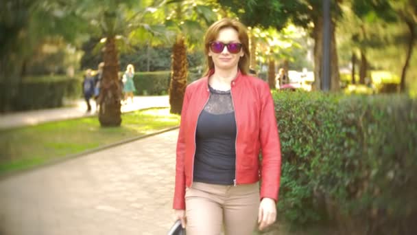 Eine Frau mit Sonnenbrille und roter Jacke schlendert an einem klaren, sonnigen Tag durch den Stadtpark. 4k, Zeitlupe — Stockvideo