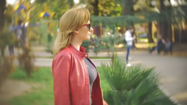 Una donna con gli occhiali da sole e una giacca rossa passeggia per il parco cittadino in una giornata di sole limpido. 4k, rallentatore — Video Stock