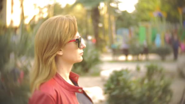 Una mujer con gafas de sol y una chaqueta roja pasea por el parque de la ciudad en un día claro y soleado. 4k, cámara lenta — Vídeo de stock